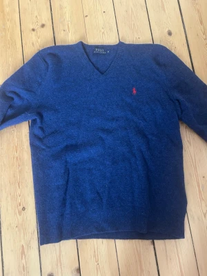 Mörkblå stickad tröja Polo Ralph Lauren - Mörkblå stickad tröja från Polo Ralph Lauren med V-ringning och klassisk röd logga broderad på bröstet. Den har också ett litet hål på ryggen men går fortfarande att använda. Tröjan har långa ärmar och ribbade muddar vid ärmslut och nederkant. Perfekt för lager på lager och stilren look. 