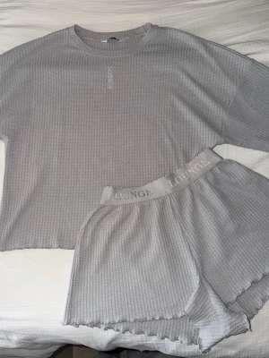 Grått ribbat loungewear-set - Mysigt loungewear-set i ljusgrått med ribbad struktur. Setet består av en tröja och matchande shorts med elastisk midja där det står 'LOUNGE'. Perfekt för chill dagar hemma.
