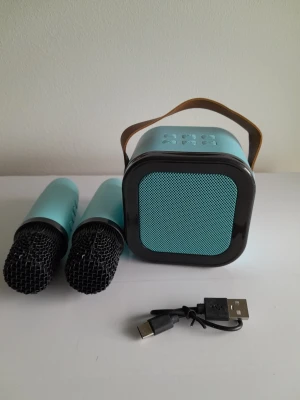 Helt nytt bärbart färgglatt RGB Bluetooth-karaokesystem med dubbla trådlösa mikrofoner - ​Ta med festen överallt med detta helt nya och extremt bärbara färgglada karaokesystem. Det är perfekt för sammankomster, fester eller solo-övningar, och denna kompakta kubhögtalare kombinerar levande visuella effekter med en kraftfull ljudupplevelse. ​Nyckelfunktioner ​Levande RGB-ljusshow: Har en dynamisk, flerfärgad LED-ljusring runt det främre gallret som ändras och pulserar för att skapa den perfekta feststämningen. ​Dubbla trådlösa mikrofoner: Levereras komplett med två matchande ljusblå t