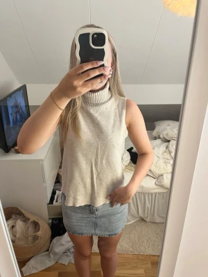 Beige ärmlös polotopp - Snygg beige ärmlös polotopp med hög krage och avslappnad passform. Perfekt att styla med kjol eller jeans för en trendig look. Materialet känns mjukt och lätt, vilket gör toppen skön att bära under varmare dagar.