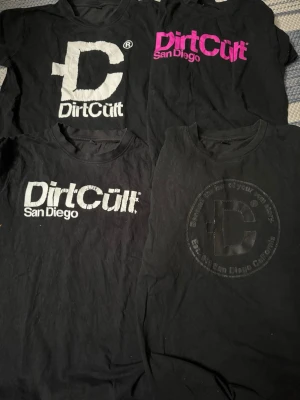 Svarta Dirt Cult t-shirts med tryck - Fyra svarta t-shirts från Dirt Cult med olika tryck i vitt, rosa och svart. Alla har rund hals och korta ärmar. Trycken varierar mellan stor logga, texten 'San Diego' och olika färgkombinationer. Perfekta för dig som gillar streetwear och grafiska prints. Det är st 146/152 på 1 T-shirt o de övriga tre är det 158/164. T-shirten är i gott skick med trycken har krakelerat 