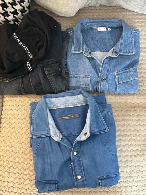 3 jeansskjortorden svarta storlek 170, den blå som är nere storlek L, den som är uppe till höger är storlek 164. - Två klassiska blå jeansskjortor i denim med knappar framtill och bröstfickor. Perfekta att styla till både t-shirt och tröja. En av skjortorna är från Dressmann.