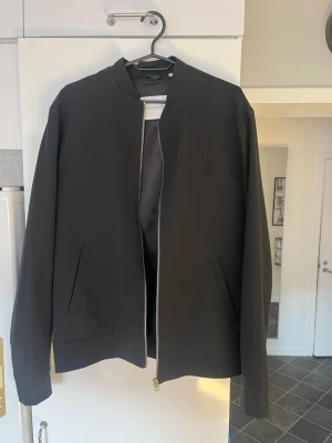 Svart bomberjacka från Les Deux - Stilren svart bomberjacka från Les Deux med minimalistisk design, silverfärgad dragkedja och två snedställda fickor framtill. Jackan har ribbade muddar och är tillverkad i ett slätt tyg som ger en clean look.