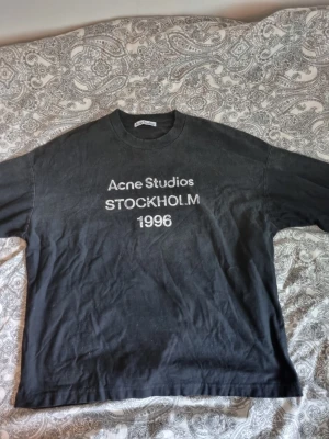 Svart långärmad tröja Acne Studios - Svart långärmad tröja från Acne Studios med tryck i vitt på bröstet där det står 'Acne Studios STOCKHOLM 1996'. Klassisk rund halsringning och avslappnad passform. Perfekt för dig som gillar minimalistisk och stilren design.