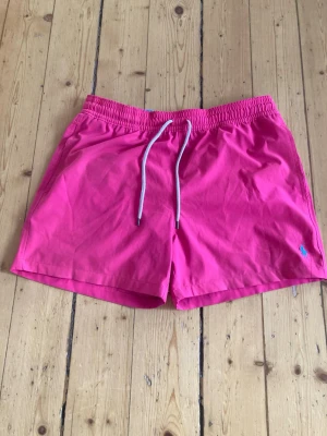 Rosa badshorts från Polo Ralph Lauren - Snygga rosa badshorts från Polo Ralph Lauren med vit snörning i midjan ock klassisk blå logga broderad på benet. Shortsen har elastisk midja och är perfekta för sommardagar vid stranden eller poolen.