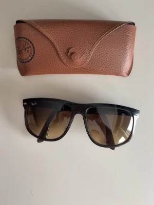 Ray-Ban - Klassiska svarta Ray-Ban Wayfarer solglasögon med bruna tonade glas. Snygg och tidlös design med Ray-Ban-logga på sidan. Kommer med ett brunt fodral i skinnimitation. Perfekta för soliga dagar och ger en cool vibe. På sista bilden ser man att knappen på fodralet har gått sönder lite men påverkar ingenting!