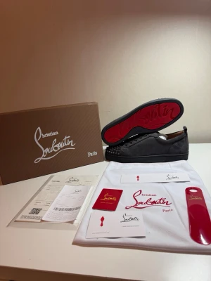 Christian Louboutin svarta sneakers - Snygga svarta sneakers från Christian Louboutin med klassisk röd sula och nitar på tån. Skorna är låga och har en ovandel i mocka samt detaljer i läder. Perfekta för dig som vill sticka ut med exklusiv stil.