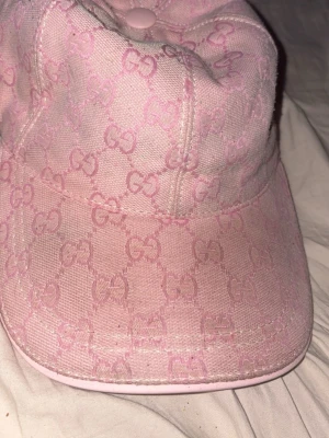 Ljusrosa keps med GG-mönster - Säljer en ljusrosa keps från Gucci med det klassiska GG-monogrammet i hela tyget. Kepsen har böjd skärm, justerbart band bak i matchande rosa och är tillverkad i ett slitstarkt tyg. Perfekt för dig som vill ha en trendig accessoar med lyxig känsla.