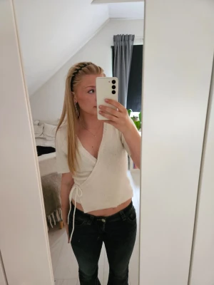 Vit omlottopp med knyt - Säljer en söt vit omlottopp med korta ärmar och knyt i sidan. Toppen är croppad och har en mjuk, stickad känsla som passar perfekt till jeans eller kjol. Enkel och stilren design som är lätt att matcha.
