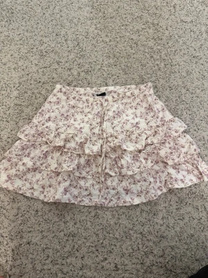Vit blommig volangkjol med snörning - Supersöt kort kjol i vitt med lila blommönster och volanger. Kjolen har snörning framtill och flera lager volanger som ger en romantisk vibe. Perfekt för varma dagar och passar till både sneakers och sandaler.