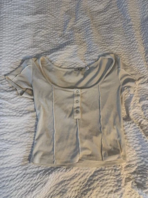 Ribbad vit topp från NA-KD - Kortärmad ribbad topp från NA-KD i vit färg. Modellen har en djup rundad urringning och knappdetaljer framtill. Tillverkad i mjukt material som sitter snyggt och bekvämt. Perfekt till jeans eller kjol.