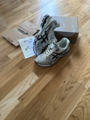 Asics gel kayano 14 - Stilrena sneakers från Asics i beige mesh med svarta detaljer och silvriga inslag. Skorna har en sportig design med tydliga Asics-loggor på sidorna och en rejäl, stötdämpande sula. Perfekta för dig som gillar en trendig och bekväm look.