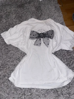 Vit t-shirt med prickig rosett - Säljer en vit t-shirt med korta ärmar och en stor svartprickig rosett i mesh på ryggen. Ovanför rosetten finns en liten text. T-shirten har en avslappnad passform och är gjord i mjukt bomullsmaterial. Perfekt för dig som vill ha något gulligt och unikt i garderoben.