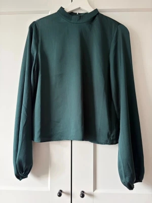 Mörkgrön blus med hög krage - Stilren mörkgrön blus med hög krage och långa ärmar. Blusen har en rak passform och knäppning i nacken med en liten öppning baktill. Materialet är lätt och ger ett elegant fall. Perfekt att matcha med både jeans och kjol.