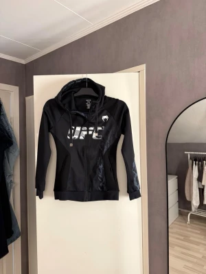 Svart UFC hoodie med dragkedja - Snygg svart hoodie med dragkedja framtill och vit UFC-logga på bröstet. Har huva med dragsko och diskreta mönsterdetaljer på ena ärmen och sidan. Tillverkad i ett sportigt material som känns bekvämt och passar perfekt för en aktiv stil.