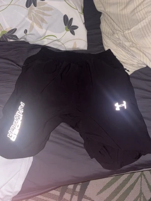 Svarta träningsshorts från Under Armour - Svarta träningsshorts från Under Armour med reflekterande logga och tryck på ena benet. Shortsen har elastisk midja med snörning och är tillverkade i ett lätt och bekvämt material som passar perfekt för träning.