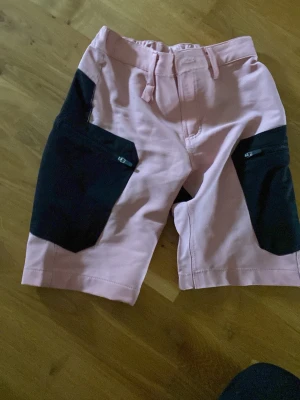 Rosa och svarta friluftshorts med fickor - Snygga friluftshorts i rosa med svarta detaljer och  fickor på framtill. Shortsen har en bekväm midja med hällor och är perfekta för en avslappnad stil. Materialet känns slitstarkt och passar bra till sommaren. Några små fläckar som syns i vissa ljus.