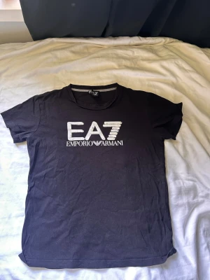 Svart EA7 Emporio Armani t-shirt - Snygg svart t-shirt från EA7 Emporio Armani med stort vitt logotryck på bröstet. Klassisk rund halsringning och korta ärmar. Perfekt till jeans eller shorts för en clean och stilren look.