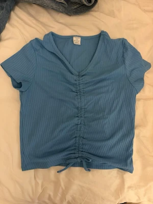 Blå ribbad topp från Lindex - Säljer en ljusblå ribbad topp från Lindex med v-ringning och korta ärmar. Toppen har snörning framtill som ger en snygg rynkeffekt och knyt längst ner. Perfekt för en trendig och avslappnad stil.