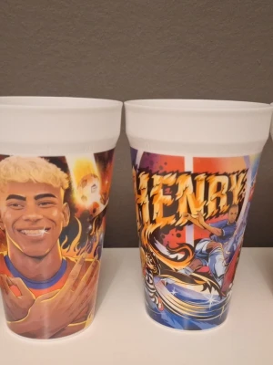 Färgglada plastmuggar med motiv - Två vita plastmuggar med färgglada tecknade motiv av fotbollsspelare och figurer. Ena muggen har texten 'HENRY' och båda har livliga illustrationer i rött, blått, gult och lila. Perfekta för dryck vid filmkväll eller gaming.