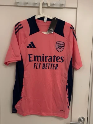 Arsenal rosa matchtröja Adidas - Säljer en rosa Arsenal matchtröja från Adidas med mörkblå detaljer och logga på bröstet. Tröjan har korta ärmar, rund hals och är tillverkad i ett lätt syntetmaterial. Perfekt för fotbollsträning eller att bära som supporter.