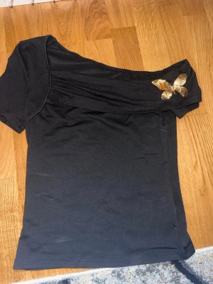 Svart oneshoulder topp  - Snygg svart oneshoulder topp med kort ärm och en guldfärgad fjärilsdetalj på axeln. Toppen har en unik skärning och är tillverkad i ett mjukt, stretchigt material som sitter bekvämt på kroppen. Perfekt för dig som vill sticka ut lite extra.