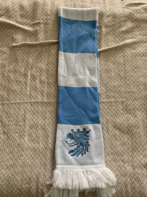 Blåvit halsduk med lejonmotiv - Snygg halsduk i blått och vitt med breda ränder och ett broderat lejonmotiv i ena änden. Halsduken har vita fransar och är perfekt för att visa ditt stöd för laget eller bara som en cool accessoar.