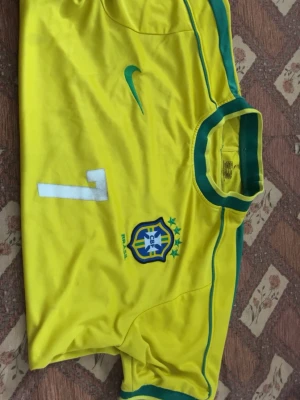 Brasilien fotbollströja Ronaldinho - Gul Brasilien fotbollströja med gröna detaljer och nummer 7. På bröstet finns landslagets emblem och Nike-logga, på ryggen står det 'Ronaldinho' och siffran 7. Tillverkad i lätt och ventilerande material, perfekt för match eller träning.