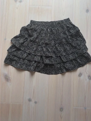 Svart blommig volangkjol - Kort svart kjol med små vita blommor och volangdetaljer. Kjolen har flera lager volanger som ger en luftig och söt look. Perfekt för dig som gillar en feminin och trendig stil.
