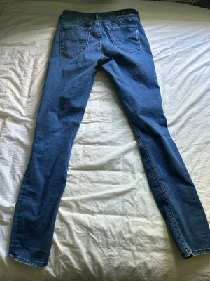 Blå skinny/slim fit jeans från Replay - Säljer ett par blå skinny jeans från Replay med klassisk femficksdesign och snygga detaljer på bakfickorna. Jeansen har en mörkblå tvätt och är tillverkade i stretchigt denimtyg för en bekväm passform. Perfekta till vardagslooken!