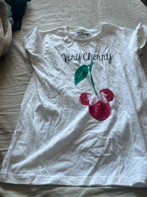 Vit Disney t-shirt med körsbärstryck - Säljer en vit t-shirt från Disney i storlek XS med ett färgglatt körsbärstryck där bären är formade som Musse Piggs huvud och texten 'Very Cherry' på bröstet. T-shirten har korta ärmar och är gjord i mjuk bomull.