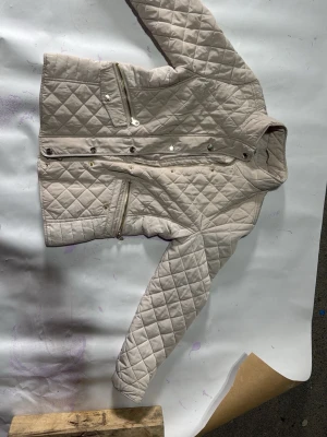 Beige quiltad jacka med dragkedjor - Snygg beige quiltad jacka med ståkrage och två snedställda dragkedjefickor framtill. Jackan har tryckknappar och är tillverkad i ett glansigt, vadderat material som ger en modern look. Perfekt för dig som gillar stilrena och tidlösa plagg. Jackan har ett litet hål på insidan.