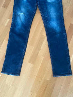Mörkblå raka jeans med slitning - Säljer ett par mörkblå jeans med raka ben och lätt slitning på vänster lår. Jeansen har klassiska fem fickor och kontrastsömmar. Materialet är denim i bomull och passformen är avslappnad men ändå snyggt figurnära.