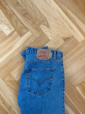 Levi's 501 blå raka jeans - Klassiska Levi's 501 jeans i ljusblå denim med raka ben och femficksmodell. Jeansen har normalhög midja, knappgylf och tydlig läderpatch bak i midjan. Perfekta för en avslappnad och tidlös stil.