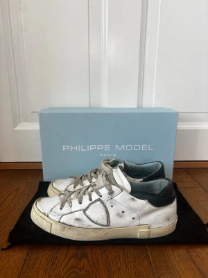 Vita Philippe Model sneakers - Snygga vita sneakers från Philippe Model med svarta detaljer på hälen och grå skosnören. Skorna har en klassisk låg siluett och är tillverkade i läder med en robust gummisula. Perfekta för dig som gillar en stilren men ändå trendig look. Lite slitna under men inget som påverkar box o allt fås med 