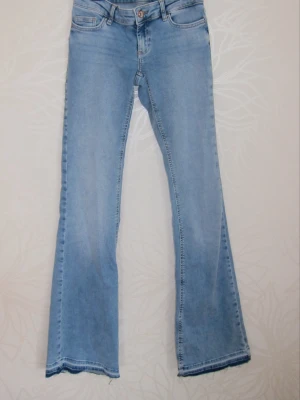 Ljusblå bootcut jeans  - Säljer ett par ljusblå bootcut jeans med klassisk femficksdesign och råa kanter nertill. Jeansen har låg midja och är tillverkade i ett mjukt denimtyg som ger en avslappnad vibe. Perfekta för dig som gillar en retro och trendig look.