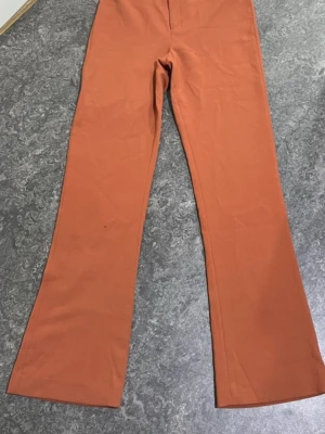 Brända orangea kostymbyxor bootcut - Snygga kostymbyxor i en bränd orange färg med bootcut-ben och hög midja. Byxorna har bälteshällor och dold stängning fram. Materialet ser ut att vara mjukt och stretchigt, perfekt för en trendig look.