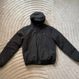 Svart quiltad vindjacka Lyle & Scott - Svart vindjacka från Lyle & Scott med huva och dragkedja. Jackan har quiltat foder, två fickor framtill med knappar och den klassiska gula loggan på bröstet. Perfekt för kyliga dagar när du vill ha en clean och stilren look.