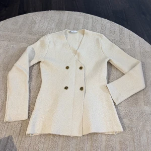 Beige kavaj med guldknappar från Zara - Snygg krämvit kavaj från Zara med dubbelknäppning och fyra guldiga knappar framtill. Jackan har en figurnära passform, v-ringning och är tillverkad i ett strukturerat tyg som känns lyxigt. Perfekt för dig som vill ha en stilren och trendig look. Endast använd 2 gånger!