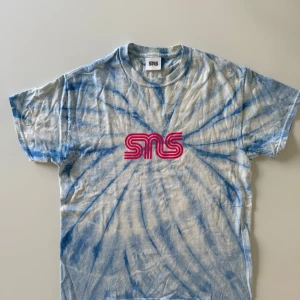 Blå tie-dye t-shirt från SNS - Snygg t-shirt från SNS med blått och vitt tie-dye mönster och ett fett rosa SNS-tryck på bröstet. Klassisk rund hals och korta ärmar, perfekt för chill dagar. Materialet är mjuk bomull som känns skönt mot huden.