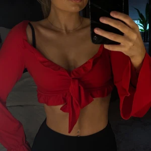 Röd croppad omlottopp med volang -  röd croppad topp med långa ärmar och volangkant. Toppen har en snygg omlottmodell med knyt framtill och v-ringning.