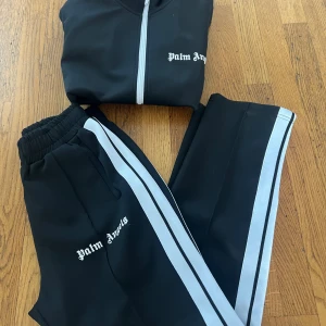 Palm Angels svarta mjukisbyxor med ränder - Svarta mjukisbyxor från Palm Angels med vita sidoränder och logga tryckt på benet. Byxorna har elastisk midja och är gjorda i polyester för en sportig vibe. Perfekta för dig som gillar streetwear och vill sticka ut med en clean look.