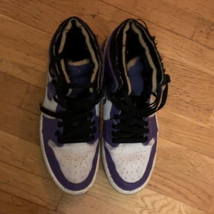 Nike Air Jordan 1 High lila och vit - Säljer ett par Nike Air Jordan 1 High sneakers i lila och vitt med svarta skosnören. Skorna har en unik design med fransade kanter och grov canvas-swoosh. Ovandelen är i skinn och textil, och sulan är vit med svarta prickar. Perfekt för dig som vill sticka ut.