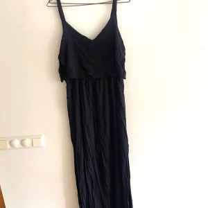 Marinblå  långklänning med smala axelband -  långklänning med smala axelband och v-ringad hals. Klänningen har en lätt rynkad struktur och ett tunt, luftigt tyg som ger en avslappnad känsla. Framtill finns en liten knytning vid bysten för extra detalj. Perfekt för varma dagar eller när du vill känna dig extra chill.