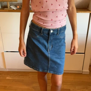 Blå jeanskjol med fickor - Snygg blå jeanskjol i klassisk modell med fem fickor, bälteshällor och knapp framtill. Kjolen är lätt A-linjeformad och slutar ovanför knäna. Perfekt till en avslappnad stil och enkel att matcha med olika toppar.