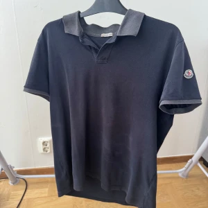 Svart Moncler pikétröja med grå krage - Svart piké från Moncler med klassisk grå krage och korta ärmar. T-shirten har Moncler-logga på vänster ärm och är gjord i bomull. Snygg och enkel design med diskreta detaljer vid krage och ärmslut. Perfekt för dig som gillar stilrena märkesplagg.