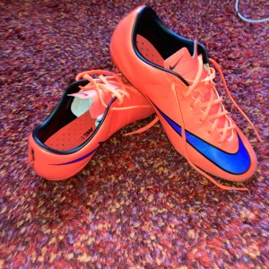 Nike orange fotbollsskor med blå swoosh - Säljer ett par riktigt snygga orangea Nike fotbollsskor med blå swoosh på sidorna. Skorna har snörning, svarta detaljer och dobbar för gräsplan. Insidan är svart och röd, och ovandelen är i syntetmaterial för bra känsla på planen.