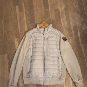 Ljusgrå quiltad jacka Parajumpers M - Snygg ljusgrå quiltad jacka från Parajumpers i storlek M. Jackan har ribbade muddar, dragkedjor på fickor och bröstficka samt patch på ärmen. Materialet är fleece och syntet, perfekt för kyliga dagar. Enkel och stilren design med ståkrage.