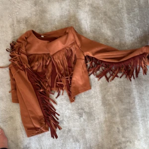 Brun mocka bomberjacka med fransar - Säljer en brun bomberjacka i mocka-look med långa fransar längs ärmar och bröst. Jackan är croppad och har en loose passform, perfekt för dig som vill sticka ut. Snygg till jeans eller klänning för en cool western vibe.