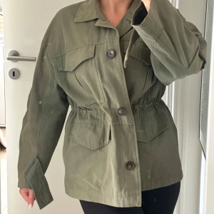Miltärjacka från Zara - Snygg grön armyjacka från Zara  med klassisk krage, stora bröstfickor och knappar framtill. Jackan har en markerad midja med resår och långa ärmar. Perfekt att slänga över en hoodie eller t-shirt för en chill look.